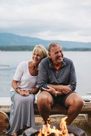 Image of an older couple sitting on a bench with a fire in the foreground and lake in the background