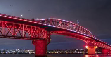 Image of the Auckland Harbour Bridge with pink and red lights at night, with the Auckland cityscape in the background