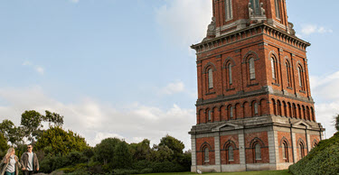 Two people walking along a path next to the historic Invercargill Water Tower, a tall red-brick structure with a domed top, set against a clear blue sky with scattered clouds.