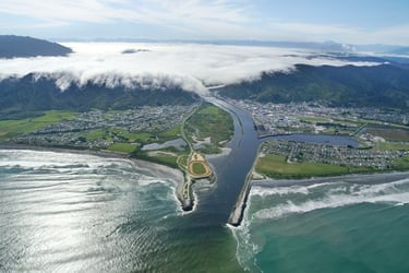 Aerial view of Greymouth with river mouth, ocean waves, town, and low clouds over green hills