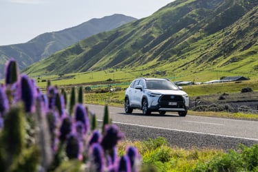 White Toyota hybrid SUV driving along a scenic road with green hills and purple flowers