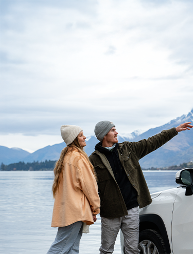 A couple in winter clothing standing by a white GO Rentals car, pointing toward snow-capped mountains beside a scenic lake in New Zealand.