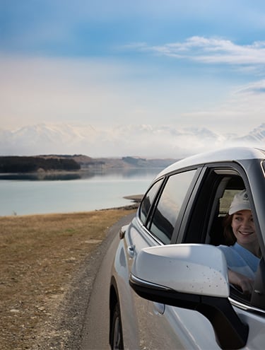 A smiling woman leans out the window of a white GO Rentals car parked beside a calm lake, with snow-capped mountains and a clear sky in the background.