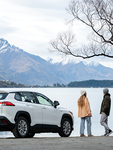 A man and woman standing by their GO Rentals hybrid car with a snowy mountain and lake backdrop.