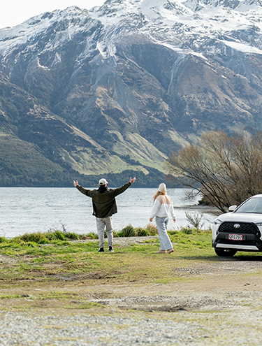 A couple stands by a lake admiring snow-capped mountains, with a white GO Rentals Hybrid SUV parked nearby.