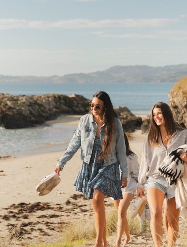 Two women walking barefoot on a beach path with towels and bags, enjoying a sunny coastal day