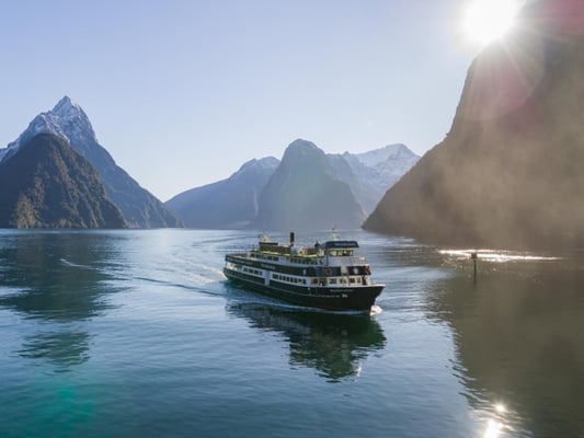 Cruise ship sailing through Milford Sound with snow-capped mountains and sunlight in the background