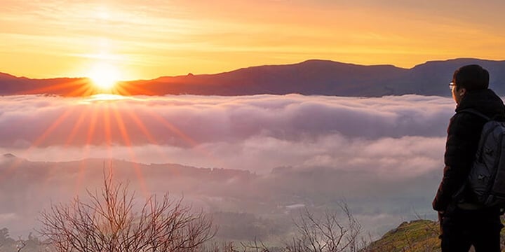 Image of a male stood on a rock at the top of a mountain overlooking a landscape covered in cloud