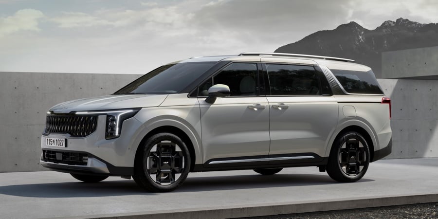 Side image of a silver Kia Carnival Hybrid people carrier parked on concrete with mountains in the background