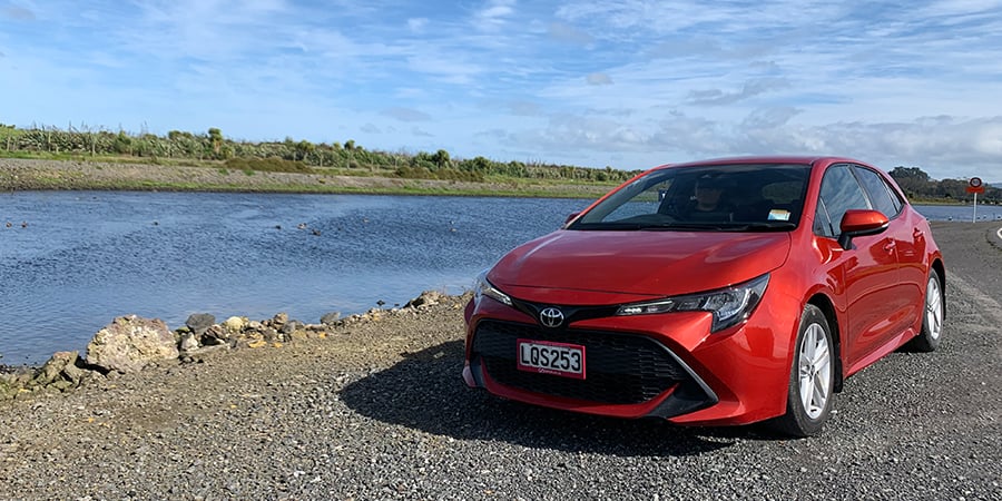 Front image of a red Toyota Corolla rental car parked up next to a river on a blue sky day