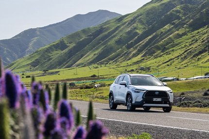 White Toyota hybrid SUV driving along a scenic road with green hills and purple flowers