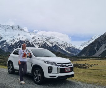 Man standing beside a white SUV with a backdrop of snow-capped mountains