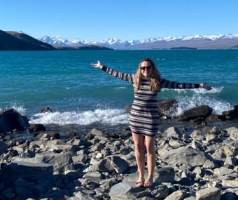 A woman standing on a rocky shore with arms open, blue lake and snowy mountains behind her