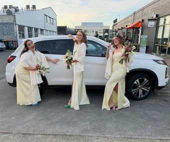 Three women in light dresses posing happily beside a white car