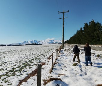 Two people walking on a snowy roadside with mountains in the background