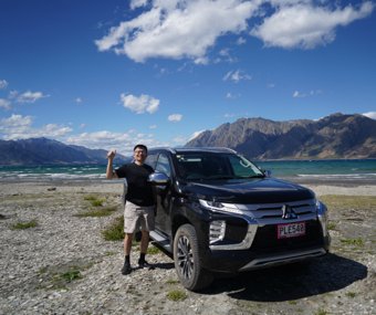 Man posing next to a black SUV with a lake and mountains in the background