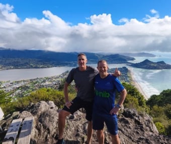 Two men posing on a mountaintop with a coastal town and ocean below