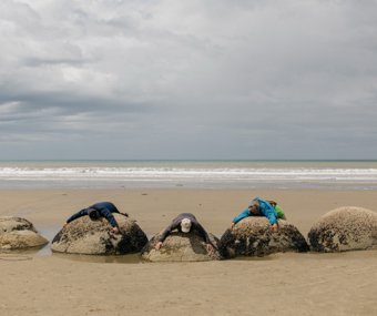 Three people lying on large round boulders at the beach under a cloudy sky