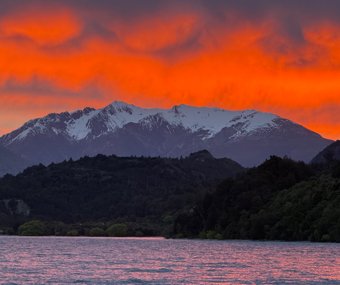 Snow-capped mountains with a dramatic orange sunset sky over a lake