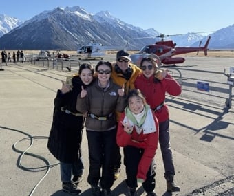 Group posing on a helipad with helicopters and mountains in the background