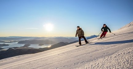 Two people skiing downhill with a mountain and lake view at sunset