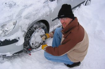 Man kneeling in the snow, installing snow chains on a car tire