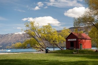 Red boathouse labeled 'Wanaka' near a lake with mountains and trees around it