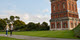 Two people walking along a path next to the historic Invercargill Water Tower, a tall red-brick structure with a domed top, set against a clear blue sky with scattered clouds