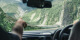 Image of a hand pointing forward from the front seat of a car as two people drive down a steep road in the South Island