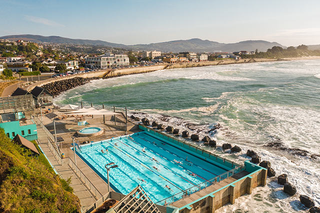 An outdoor swimming pool on the water front, the bay curves off into the distance