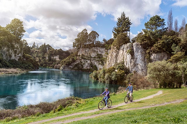 2 people cycling down a track beside a clear, blue river 