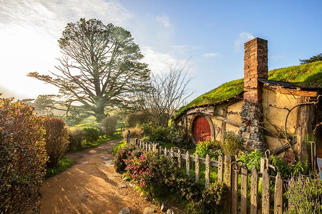 The front of a Hobbit House with a round red door and brick chimney in Hobbiton