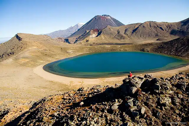 A man sat atop a rock near a sulphuric lake with mountain range in the background