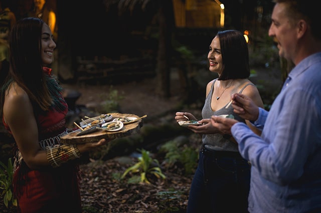 a couple eating traditionally prepared appetisers served by maori women in red dress