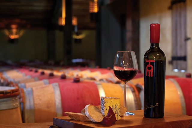 a bottle and glass of red wine with cheese in front of wine barrels 