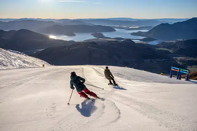 A skier and snowboarder going down the slopes towards mountains and lakes in the distance