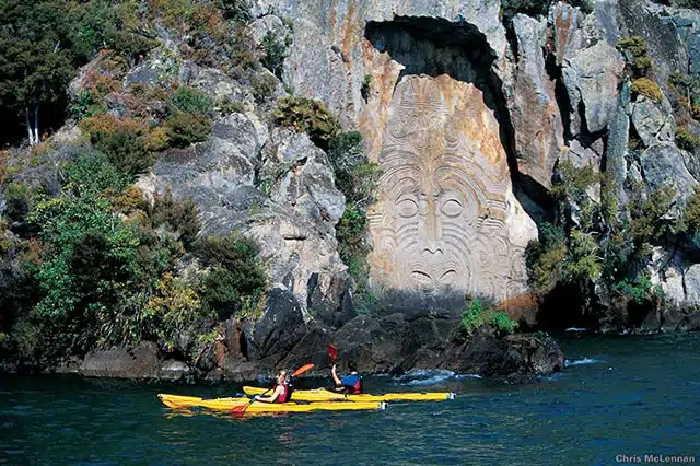 2 yellow kayaks in front of a large Maori carving that has been etched into the cliff face beside Lake Taupo