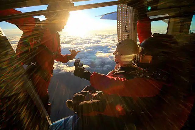 3 skydivers in orange jumpsuits preparing to jump from a plane above the clouds