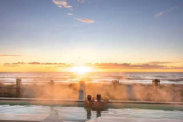 A couple with Martini glasses looking over the edge of a hot pool out towards the sun setting over the ocean.