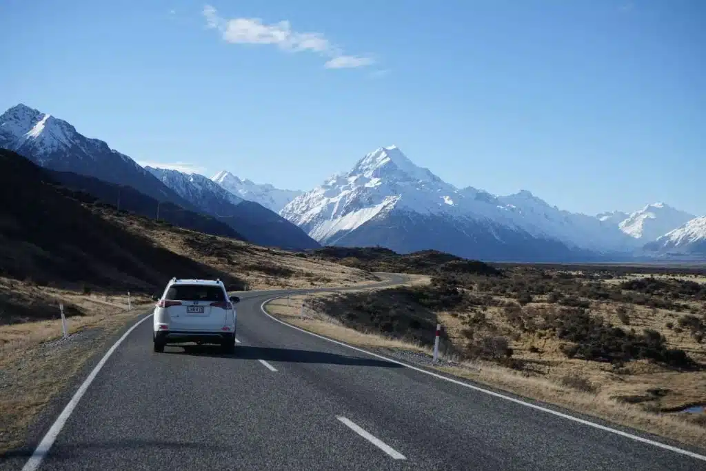 white car driving on left-hand side of the road in new zealand with mountain landscape background